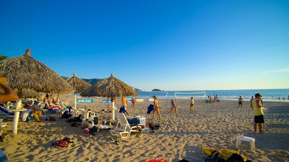 A beach with thatched umbrellas, sun loungers, and people playing volleyball.