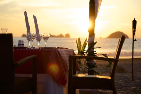 A table set for a meal on a beach with the sun setting in the background.