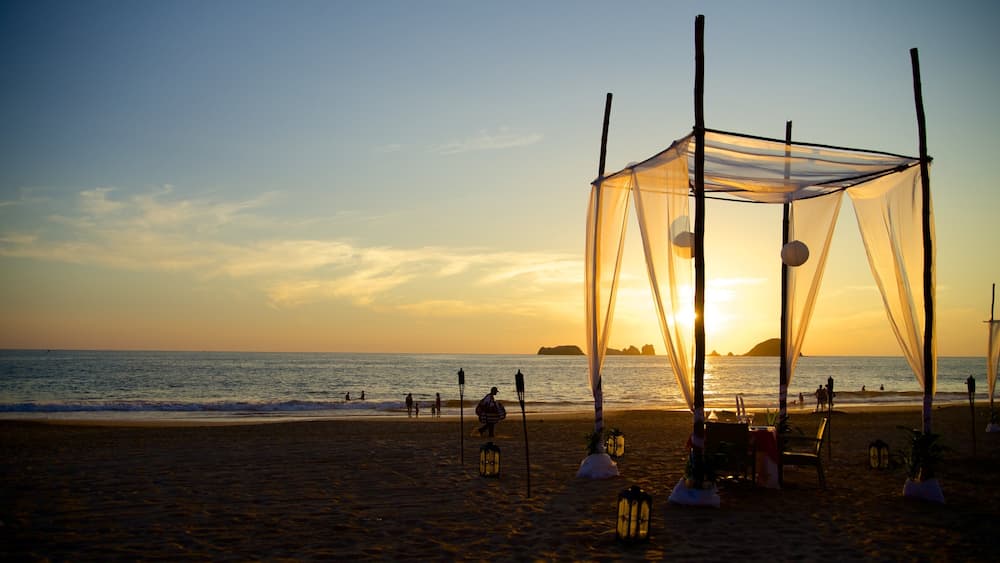 A beach with a canopy structure, lanterns, and people enjoying the sunset.