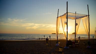 Una playa con una estructura tipo cabaña, faroles y gente disfrutando del atardecer.