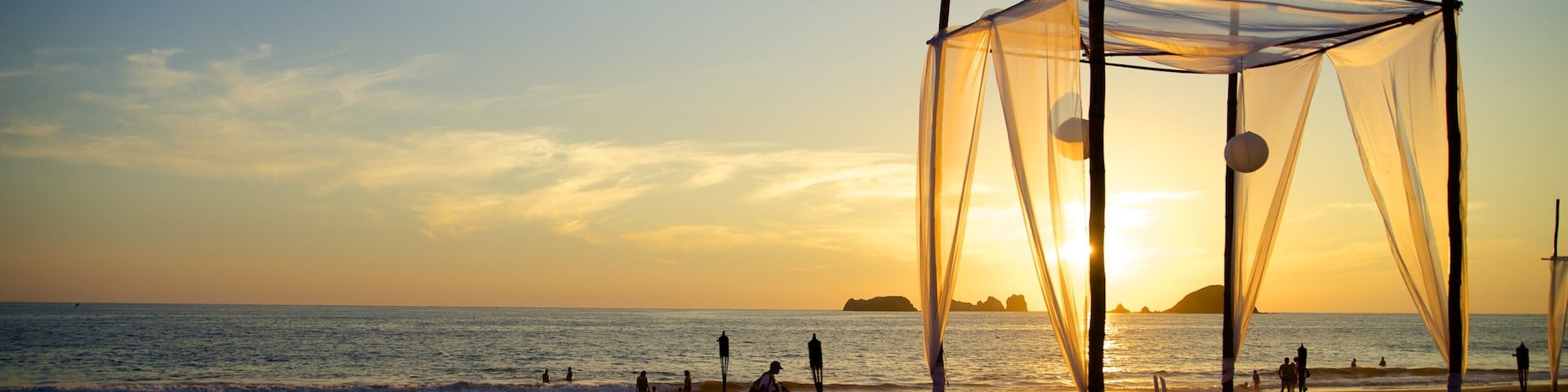 Uma praia com estrutura de toldo, lanternas e pessoas apreciando o pôr do sol.