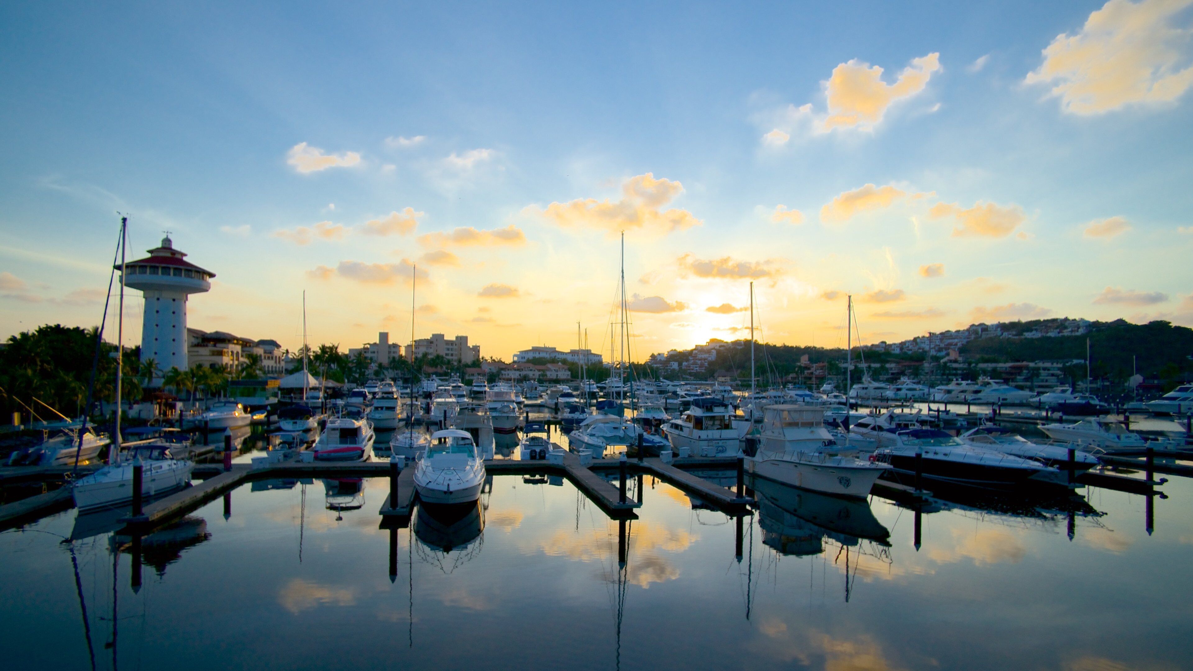 A marina with numerous boats docked, a lighthouse, and a sunset in the background.