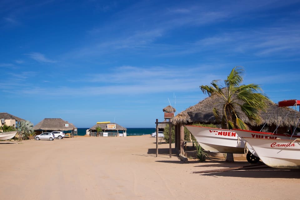 A beach with thatched-roof huts, a white boat named 'Camita', and a clear blue sky.