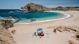 A beach with clear turquoise waters, a rocky outcrop, and a few boats.