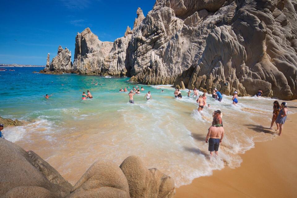Een strand met helder turquoise water, rotsachtige kliffen en mensen die zwemmen en over het strand lopen.