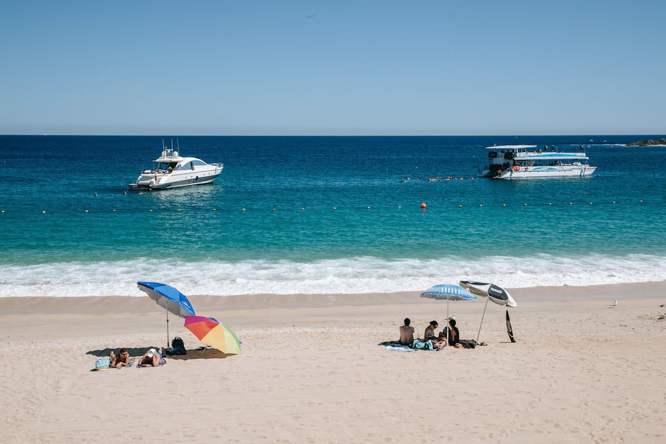 A beach with a boat in the water, people sunbathing, and a clear blue sky.