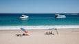 Une plage avec un bateau sur l’eau, des gens qui prennent un bain de soleil et un ciel bleu dégagé.