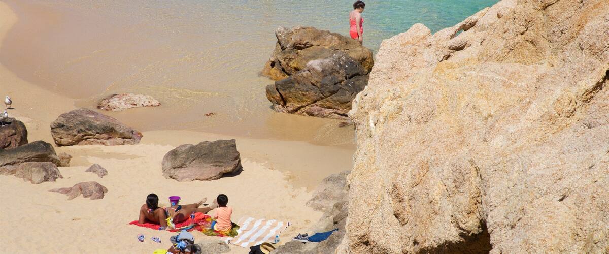 Una playa con agua turquesa cristalina, gente nadando y tomando el sol, y grandes rocas en primer plano.