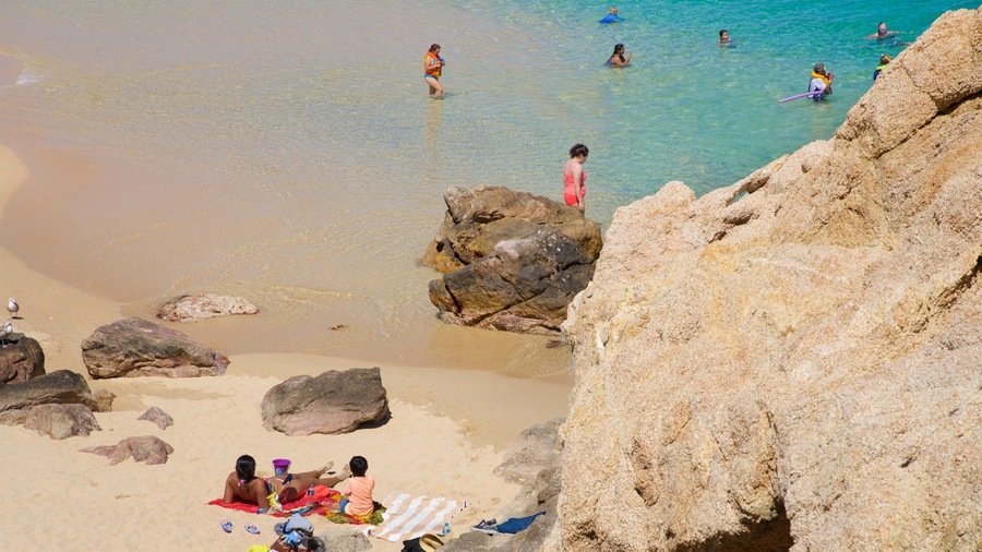 Ein Strand mit klarem, türkisfarbenem Wasser, Menschen beim Schwimmen und Sonnenbaden, und großen Felsen im Vordergrund.