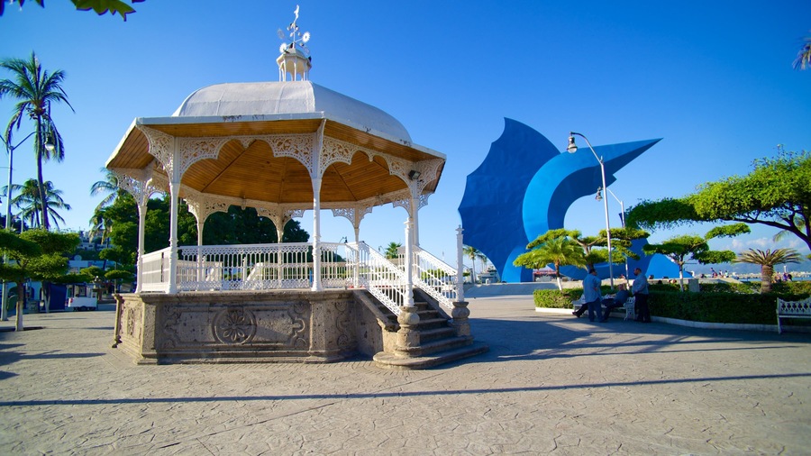 Un pabellón con elaborada celosía blanca y techo abovedado, frente a un fondo de palmeras y una gran escultura azul.