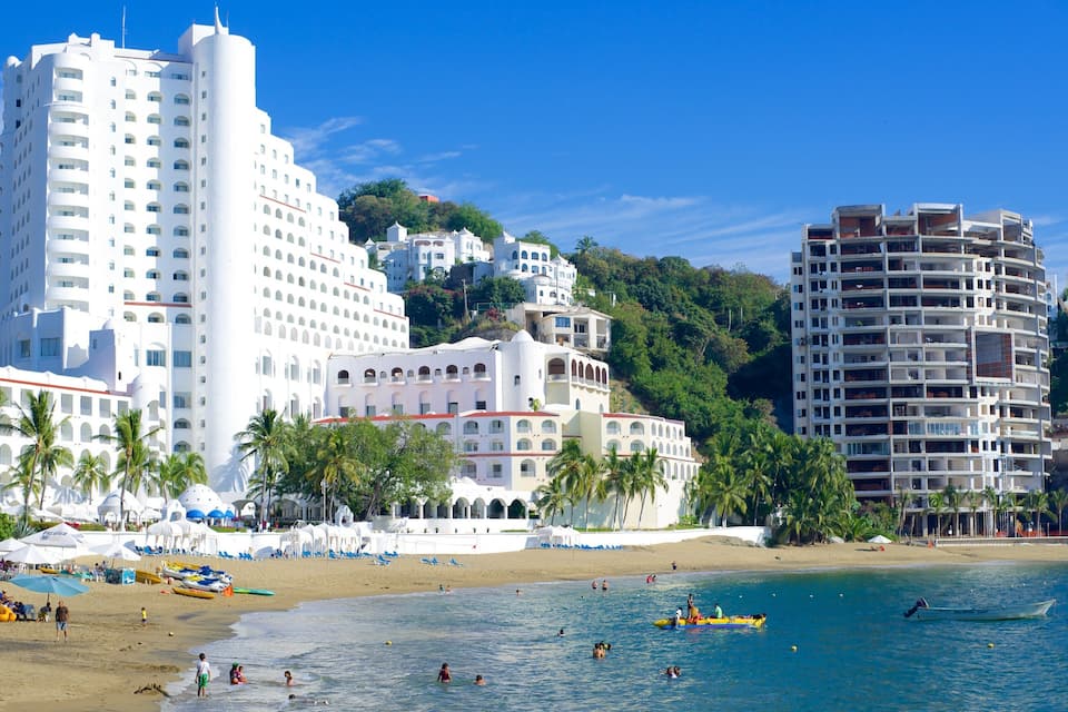 A beach with palm trees, a sandy shore, and white buildings.