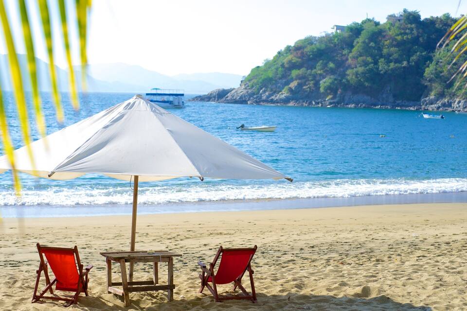 Una playa con una sombrilla blanca, dos sillas de playa rojas, una mesa de madera y un bote en el agua.