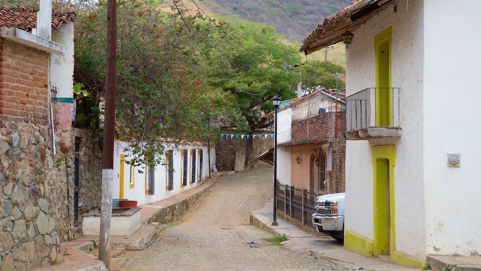 A narrow cobblestone street flanked by white buildings with yellow accents, a parked truck, and a hillside in the background.