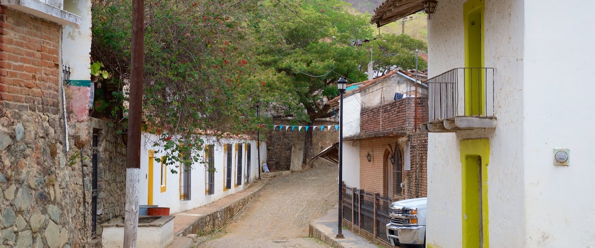 A narrow cobblestone street flanked by white buildings with yellow accents, a parked truck, and a hillside in the background.