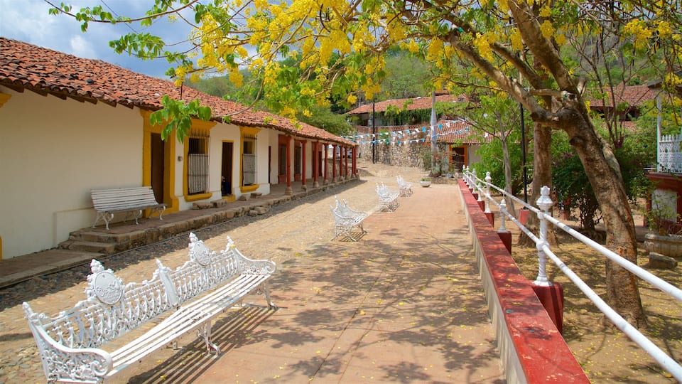 A cobblestone street lined with yellow buildings, white benches, and a stone wall with a white railing.