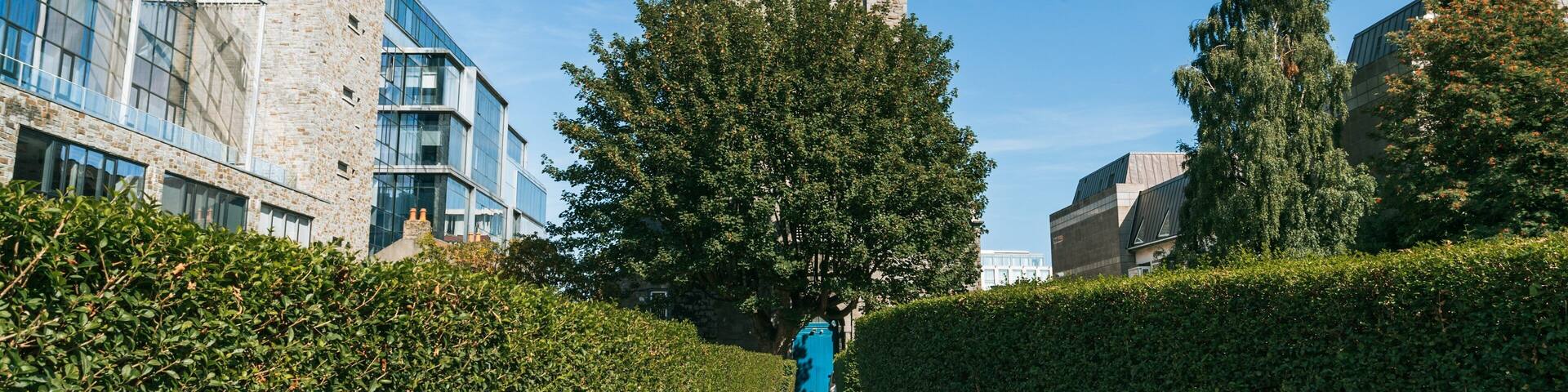 A historic stone tower, modern buildings, and a well-maintained hedge pathway.