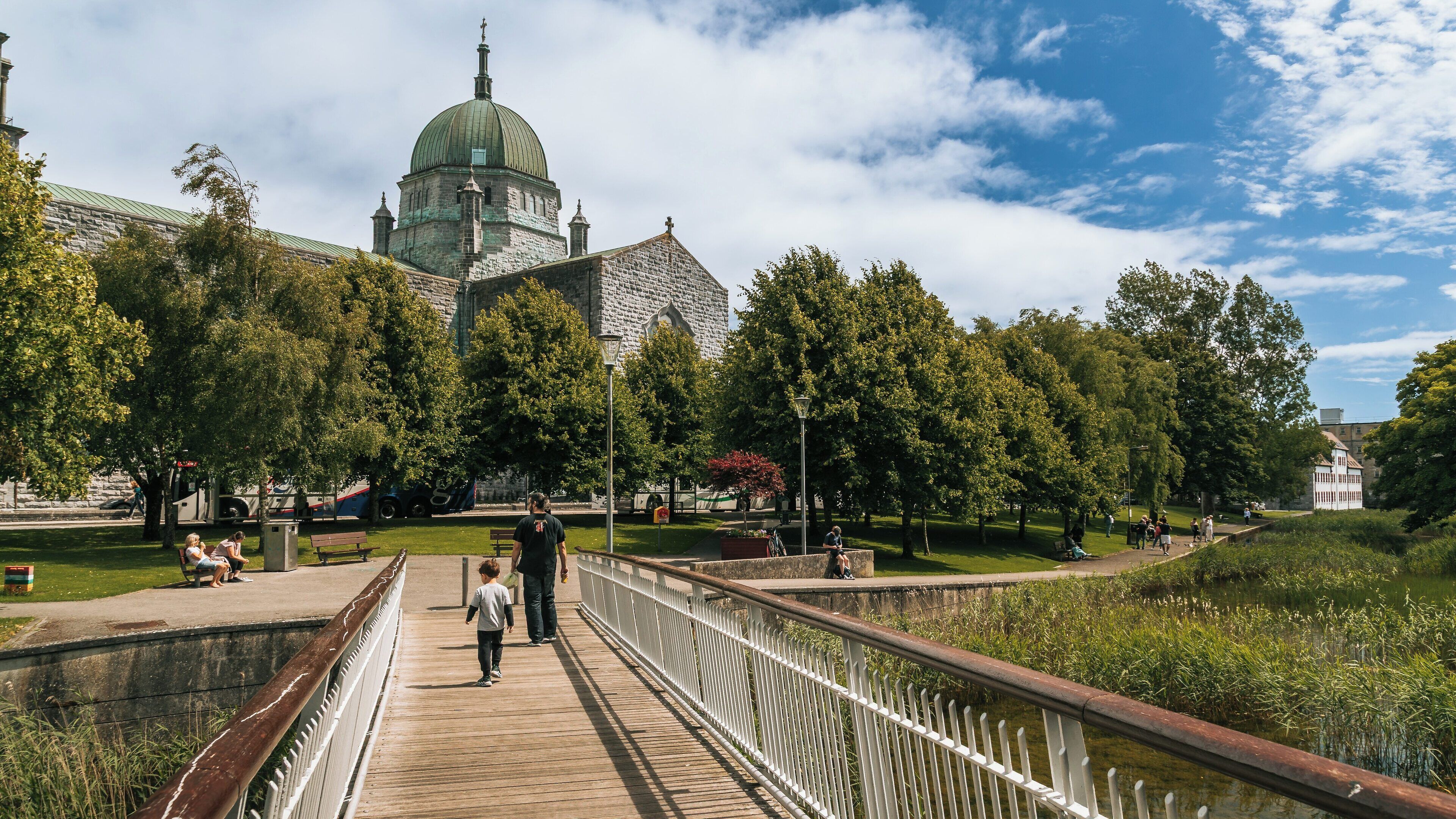 Galway Cathedral stands majestically as visitors stroll over the wooden bridge near the River Corrib on a sunny day in Galway City Centre, Ireland