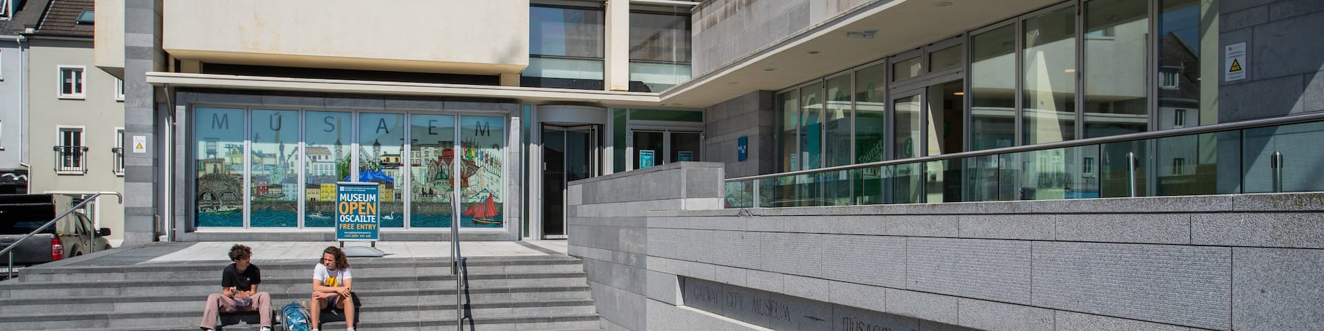 A modern building with a glass facade, a sign for the Museum of the City of Galway, and a stone wall with inscriptions.