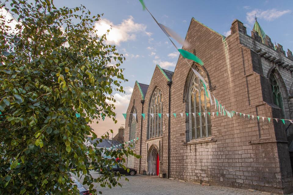 En historisk kyrka med spetsbågiga valv och en röd dörr, omgiven av träd och en klar himmel.