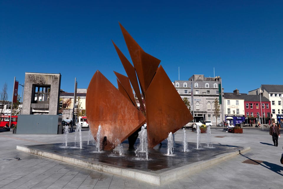 Una scultura moderna con getti d'acqua in una piazza pubblica, circondata da edifici e un cielo azzurro.