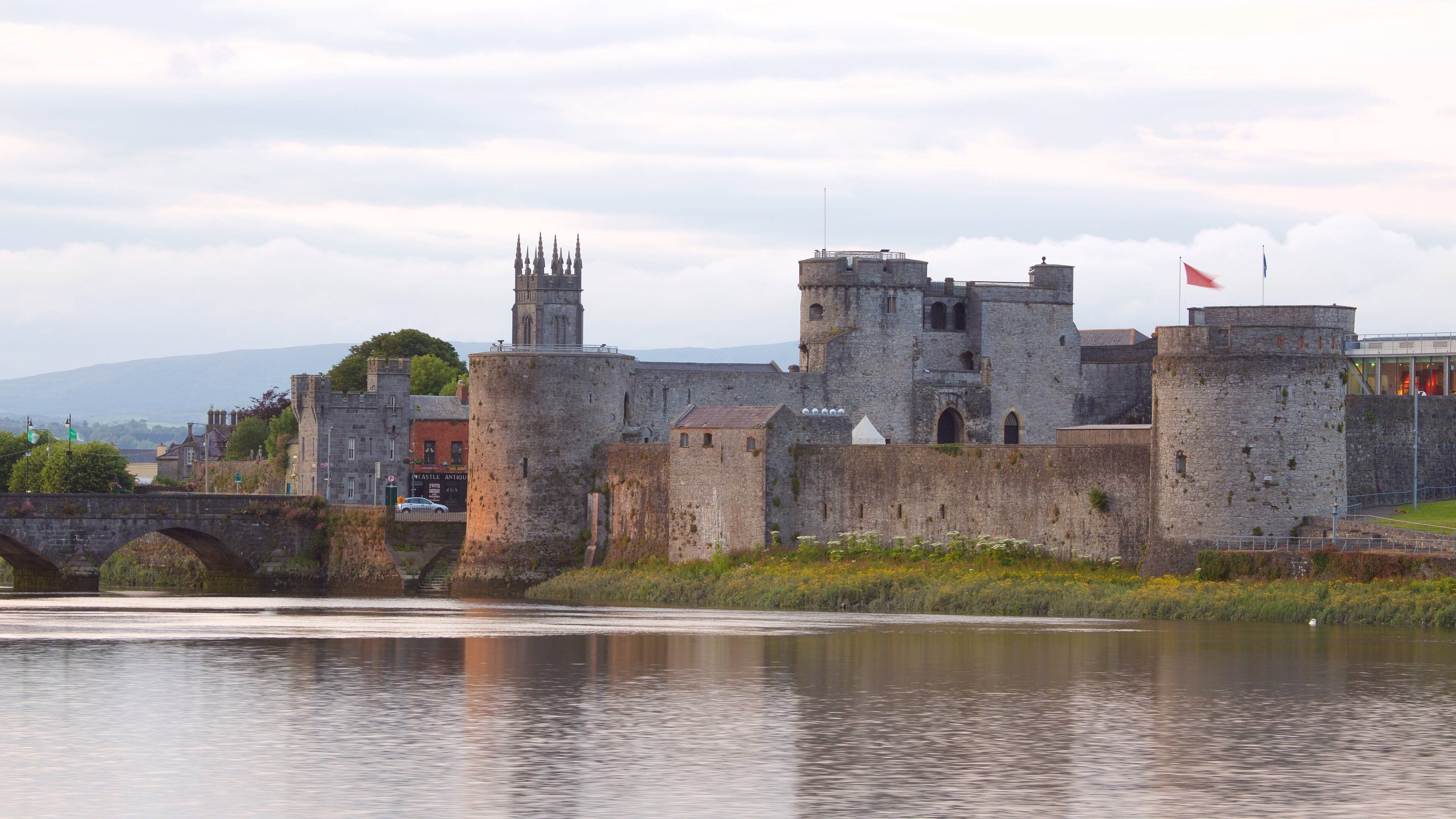 A historic castle with a bridge over a river, surrounded by greenery and a cloudy sky.