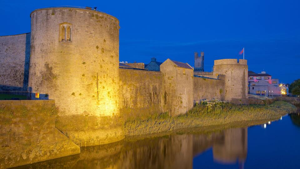 Una muralla de piedra histórica con un río en primer plano, una torre y un edificio con ventanas iluminadas al fondo.