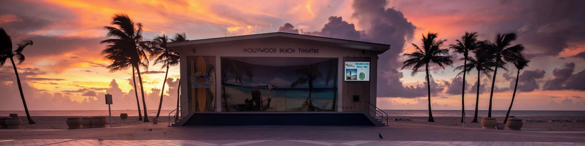 A man sitting on a tiled surface facing the Hollywood Beach Theatre with a sunset in the background.