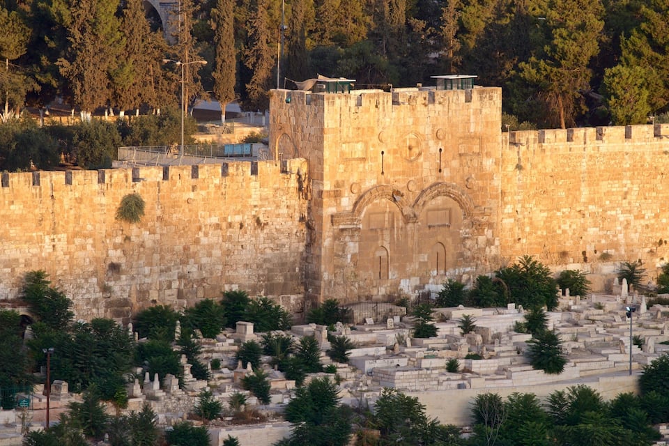 Un mur de pedra històric amb una entrada apuntada, envoltat d'un cementiri ple de tombes i arbres.