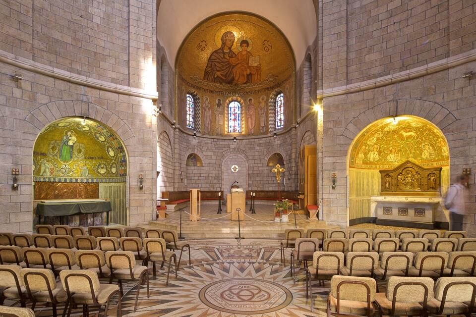 Interno di una chiesa con un altare centrale, file di bancali e mosaici decorativi sulle pareti.