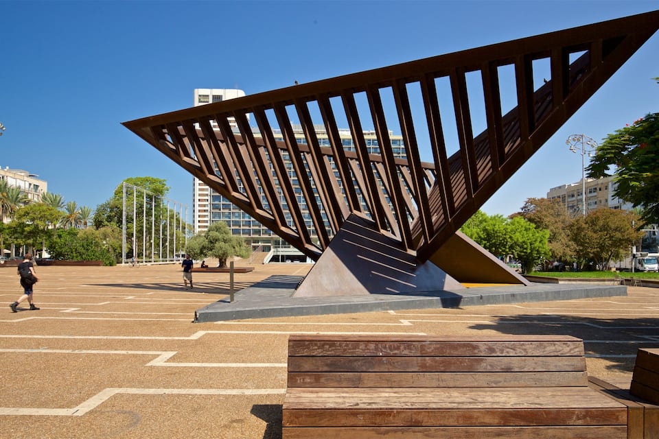 A modern sculpture in an open plaza with a clear blue sky, palm trees, and urban buildings in the background.