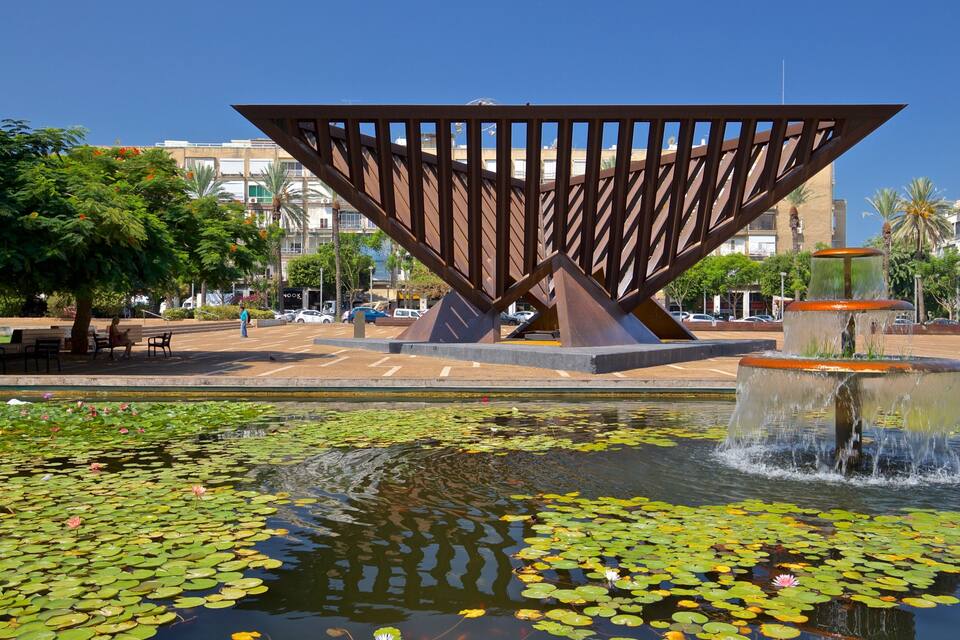 A modern sculpture in a public square with a fountain and lily pads in a pond.