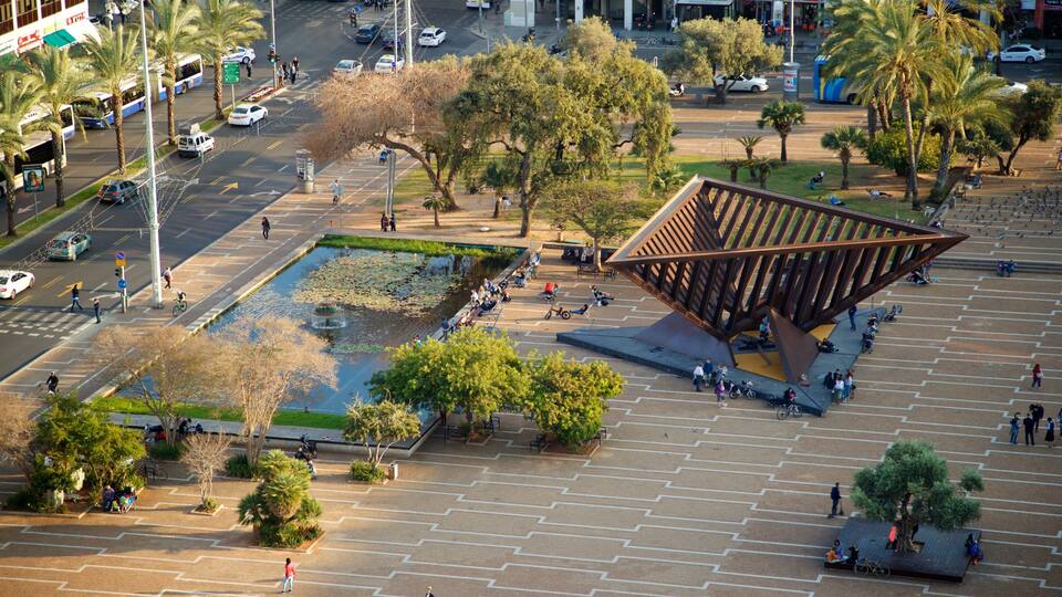 A public square with a modern sculpture, a fountain, and surrounding trees.