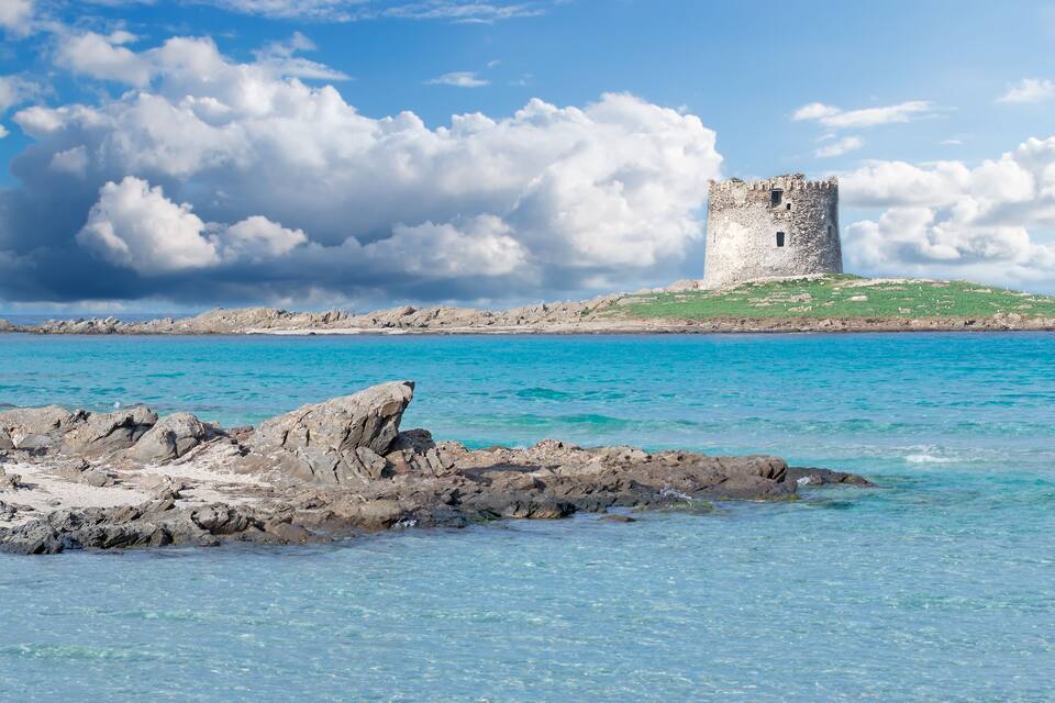 A coastal castle on a rocky outcrop with clear turquoise waters and a cloudy sky.