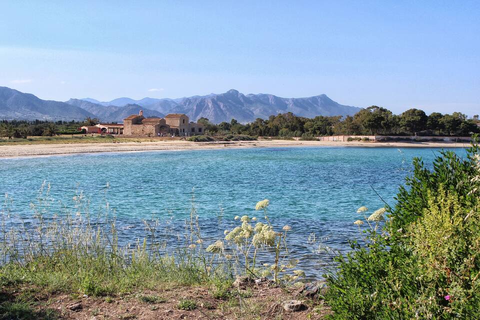 A coastal landscape with clear blue waters, a sandy beach, and mountains in the background.