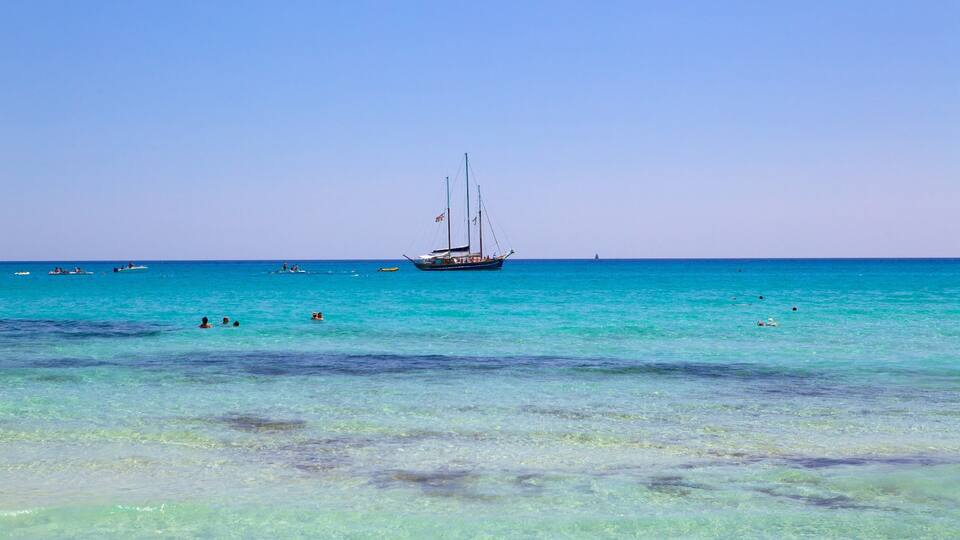 A clear turquoise sea with a sailboat in the distance and several smaller boats.