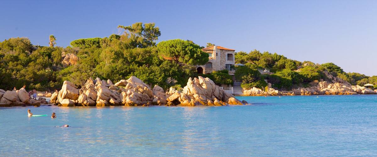 A coastal scene with clear blue water, rocky outcrops, and a building on a hill.