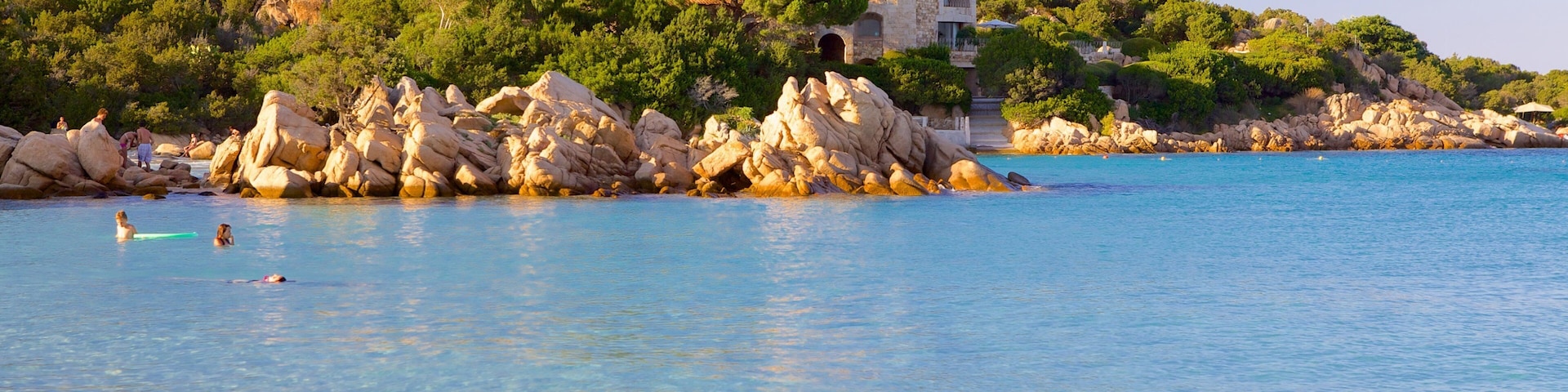 A coastal scene with clear blue water, rocky outcrops, and a building on a hill.