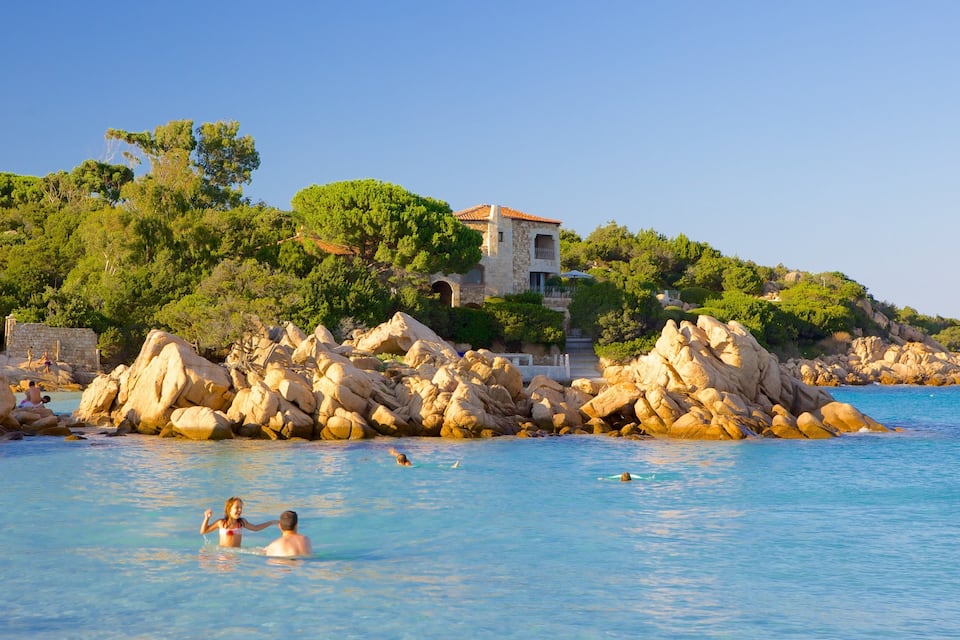 A coastal scene with clear blue water, rocky formations, and a villa on a hillside.