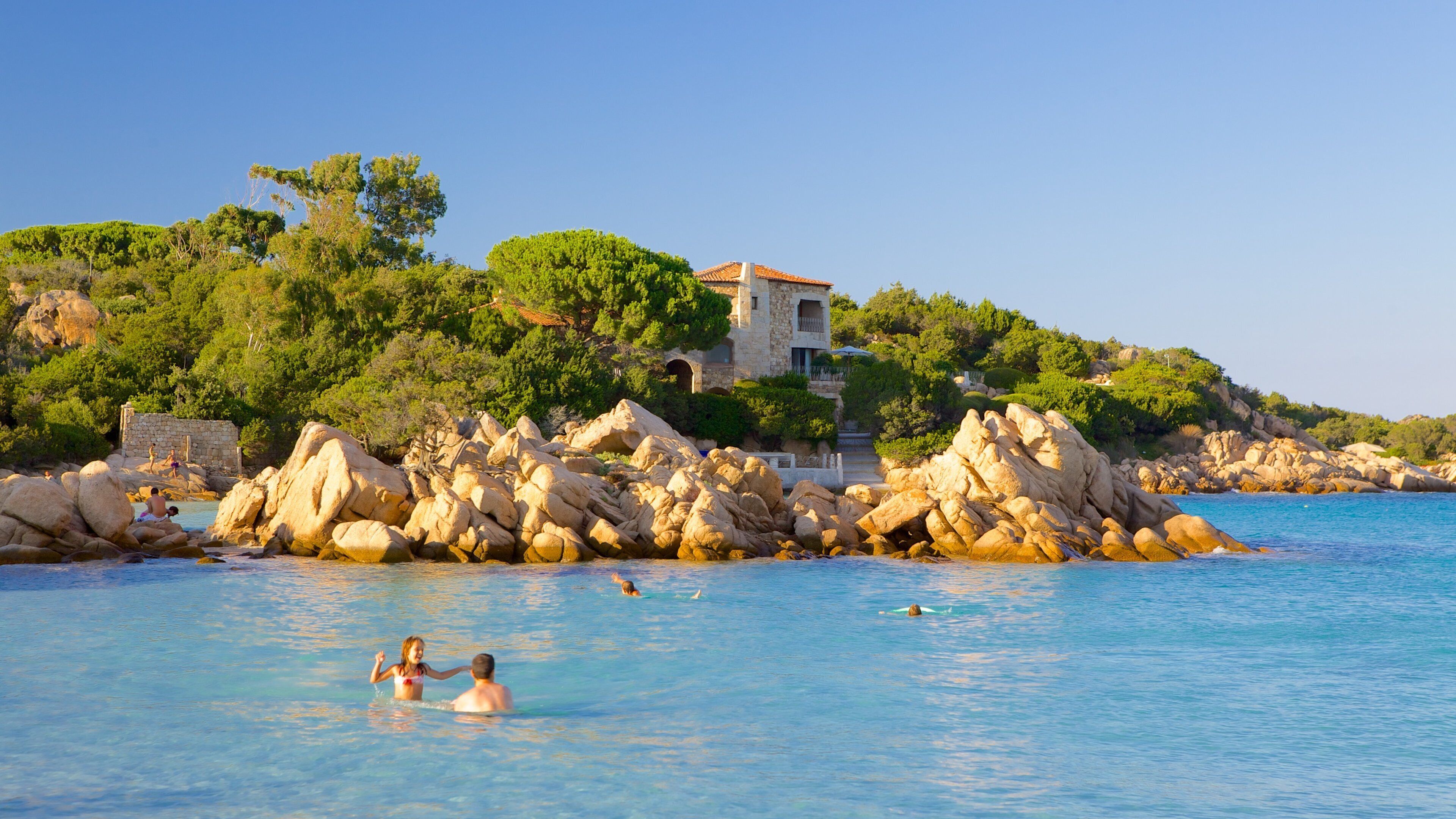 A coastal scene with clear blue water, rocky formations, and a villa on a hillside.