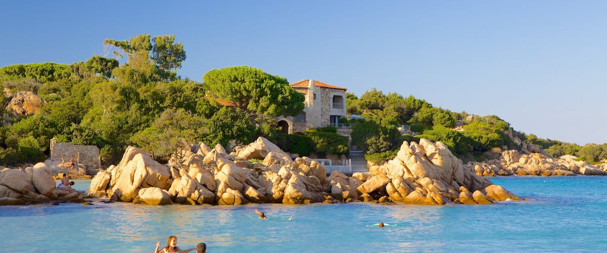 A coastal scene with clear blue water, rocky formations, and a villa on a hillside.