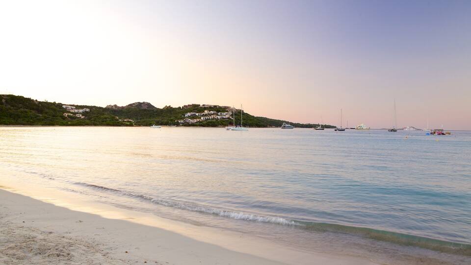 A beach with calm waters, boats, and houses on a hillside.