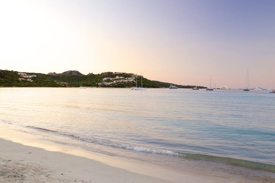 A beach with calm waters, boats, and houses on a hillside.