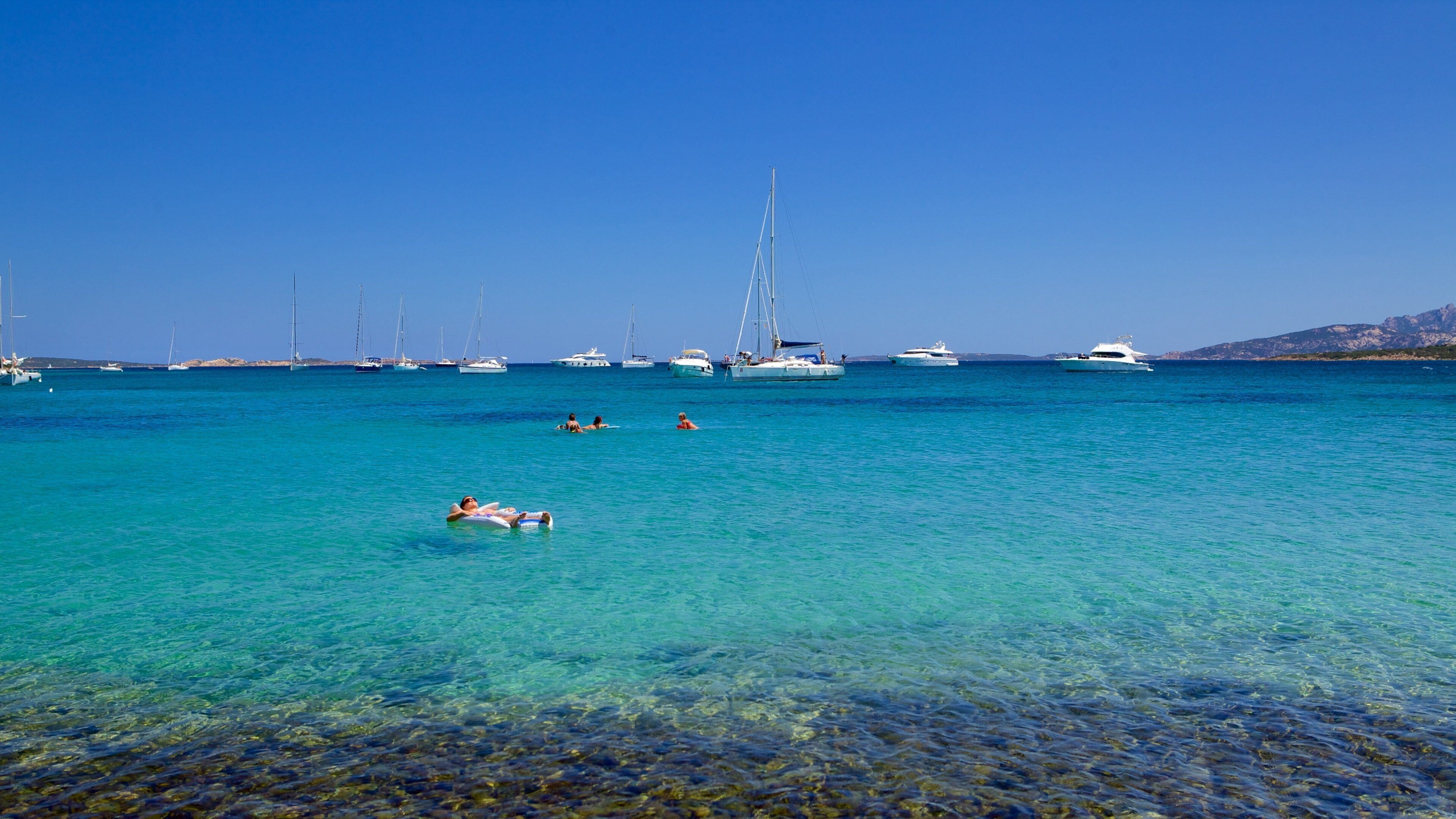 A clear turquoise sea with boats and people swimming.
