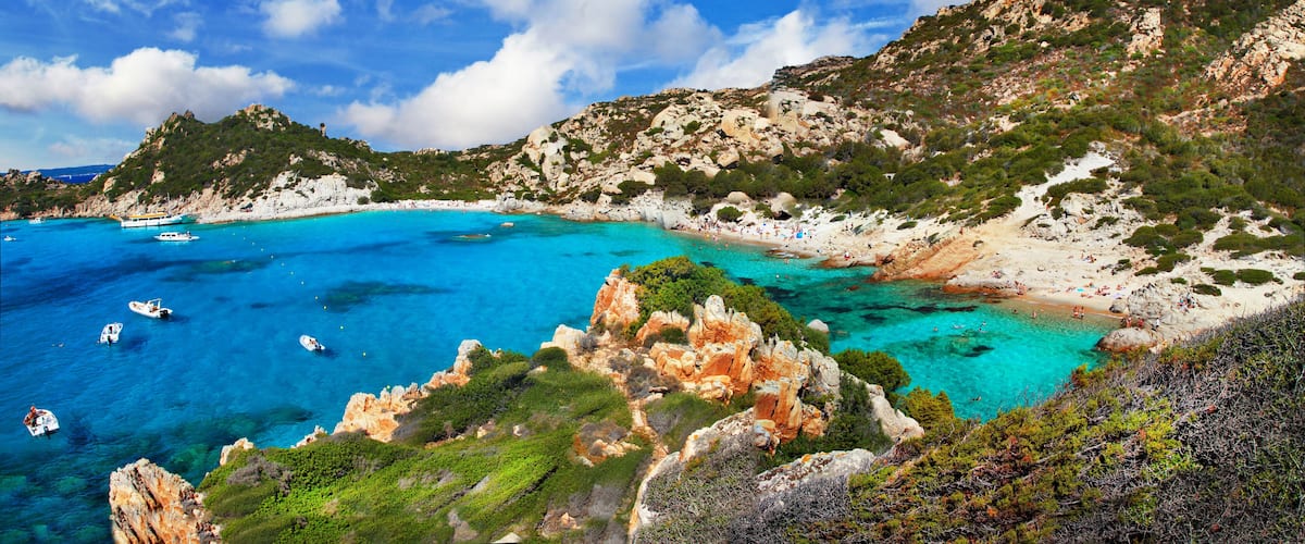 A coastal landscape with clear turquoise waters, rocky cliffs, and a beach populated with people.