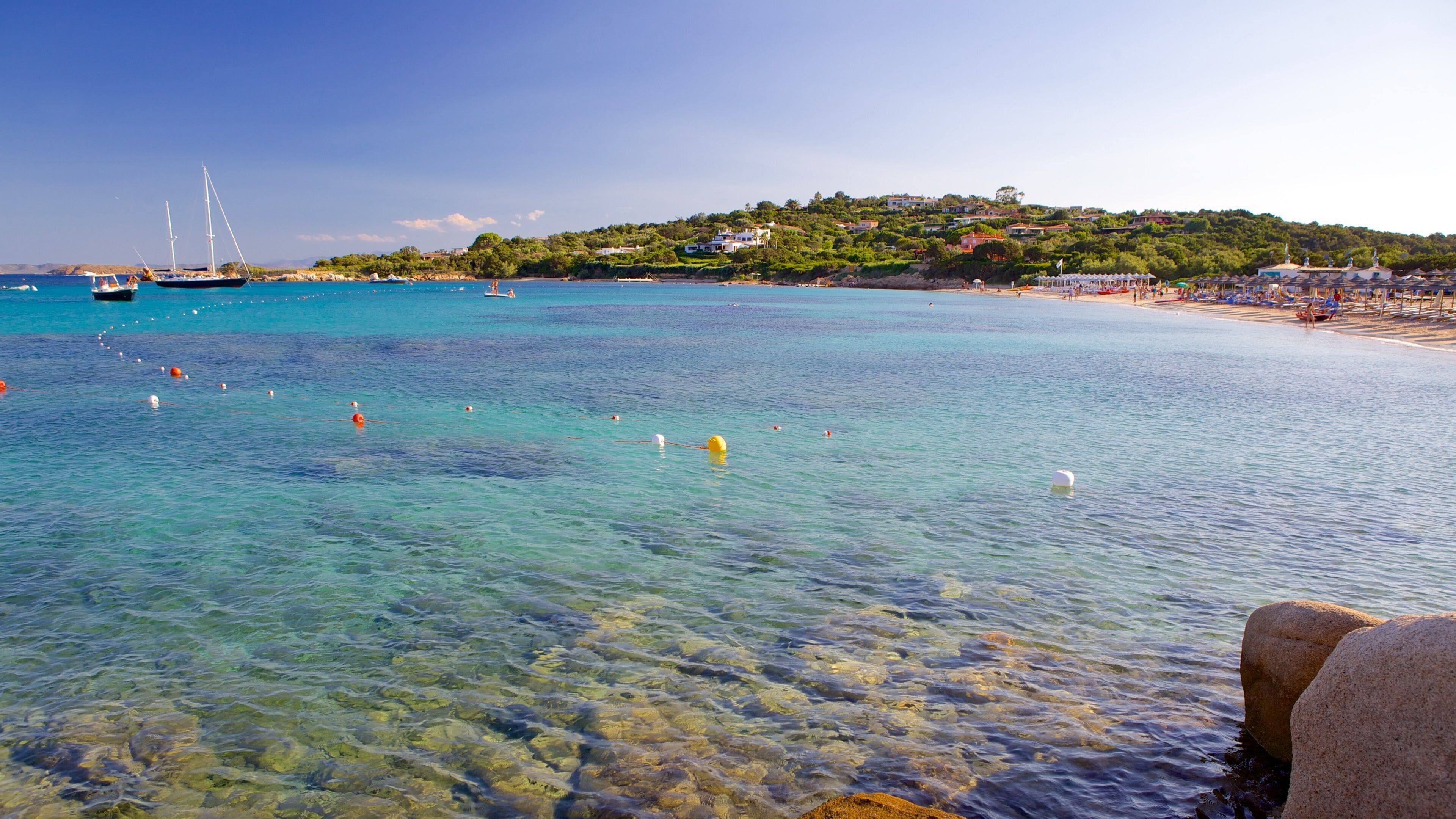 A clear turquoise sea with boats, a rocky shoreline, and a hillside with buildings.