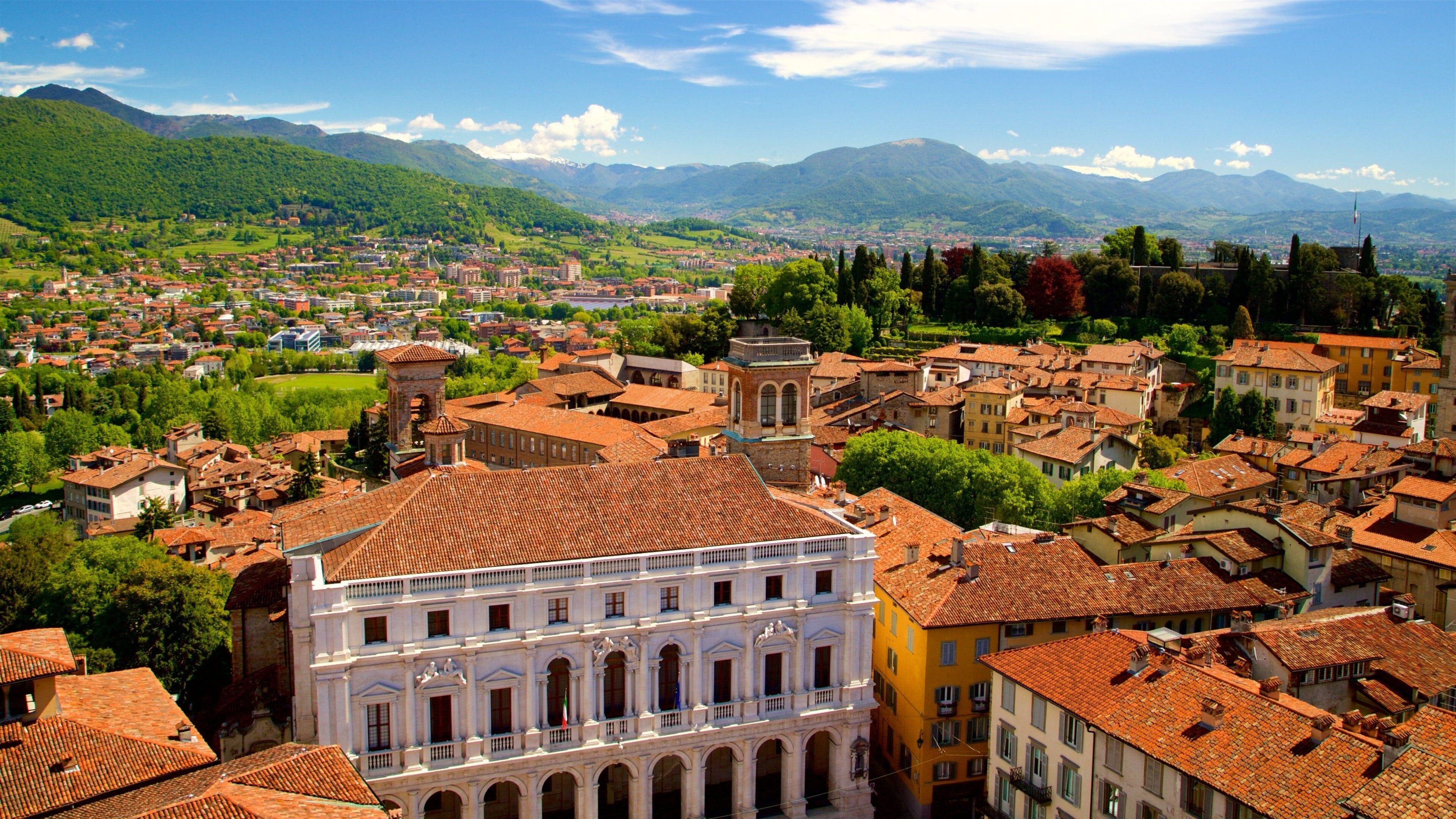 A historic town with terracotta-roofed buildings, a prominent white building with arches, and lush green hills in the background.
