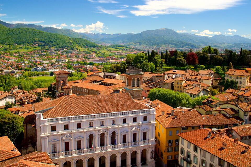 A historic town with terracotta-roofed buildings, a prominent white building with arches, and lush green hills in the background.