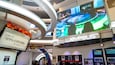 Un espace de divertissement intérieur moderne, doté d’un grand écran diffusant un jeu de course, d’une enseigne indiquant « Joypolis » et de divers posters et éléments de décoration.