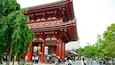 A traditional Japanese gate with red pillars and intricate carvings, surrounded by lush greenery and people.