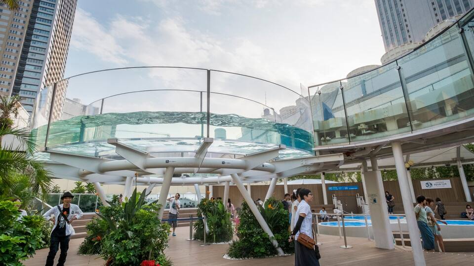Ein moderner Stadtplatz mit einer Glasarchitektur, umgeben von hohen Gebäuden und Grünflächen.